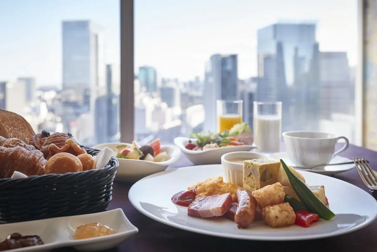 Breakfast spread on a table with a city skyline view, including bread, salad, fruit, assorted breakfast items, juice, coffee, and tea.