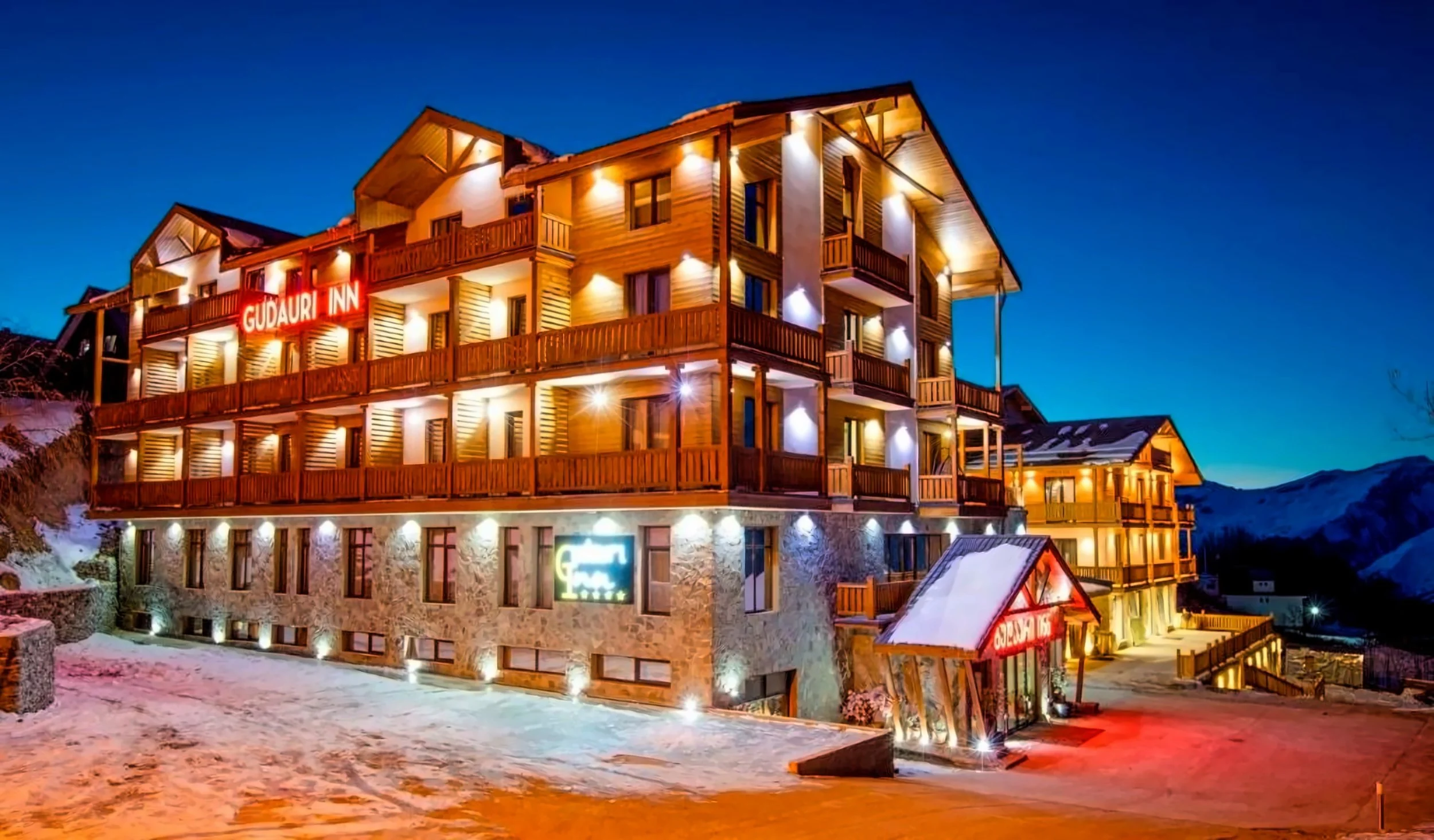 A multi-story hotel with wooden balconies illuminated by exterior lights at night, snow on the ground, and a mountain background. The hotel has signs reading 'Gudauri Inn' and 'Guest House'.