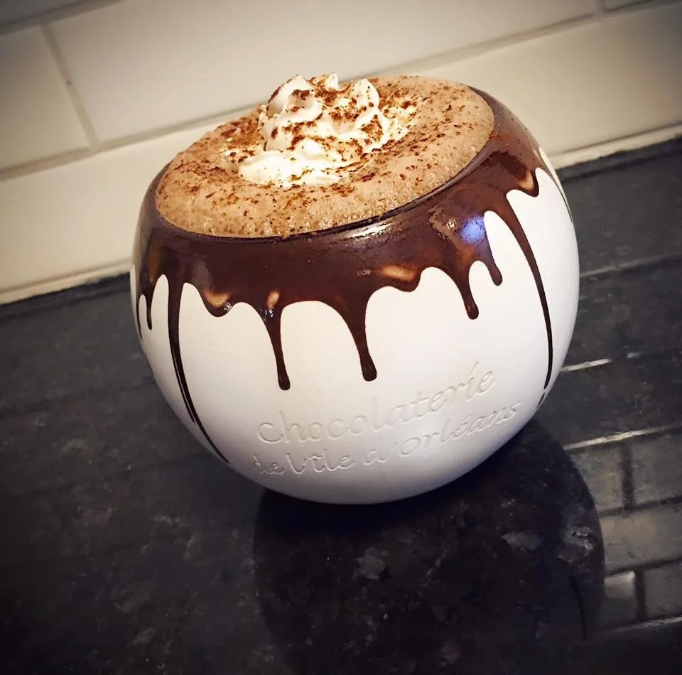 A white ceramic cup with the words 'chocolatote be vite a Orleans' written embossed on it, filled with hot chocolate topped with whipped cream and cocoa powder, and decorated with chocolate drizzle around the rim.