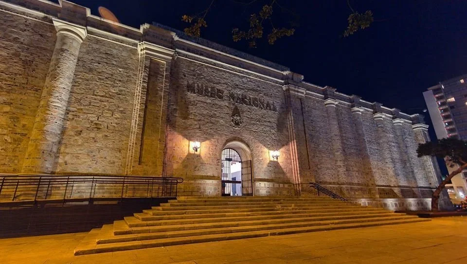 Night view of the front entrance of a historic building, labeled 'Museo Nacional,' with stone steps and iron railings, lit by exterior lights.