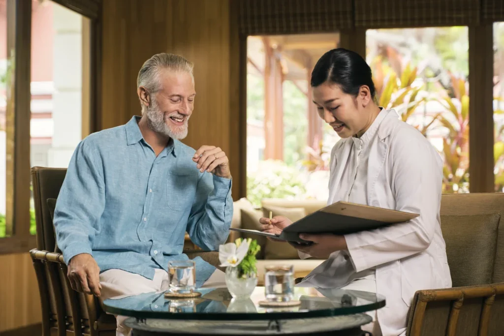 A senior man and a woman in a white coat smiling and talking at a dining table, with a woman in a white jacket looking at a menu or notepad in a cozy, well-lit restaurant with large windows and plants outside.
