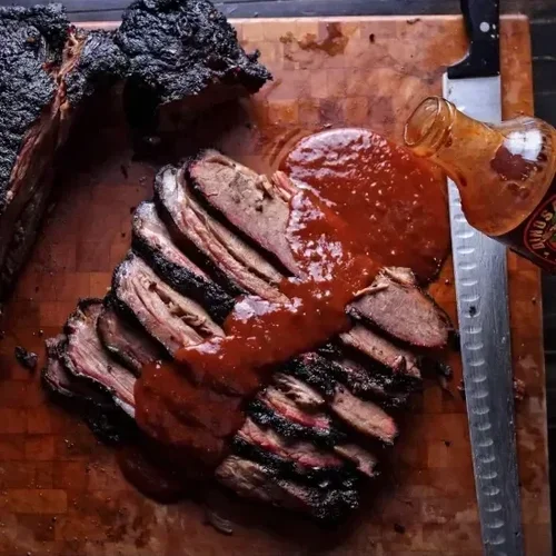 Sliced smoked brisket with barbecue sauce on a wooden cutting board, with a bottle of barbecue sauce and a knife.