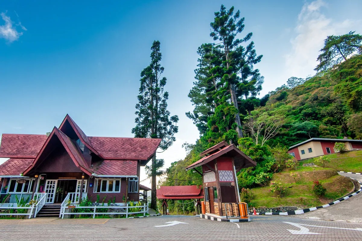 A house with a red-tiled roof, a small gatehouse, and tall trees on a hillside with other buildings in the background.