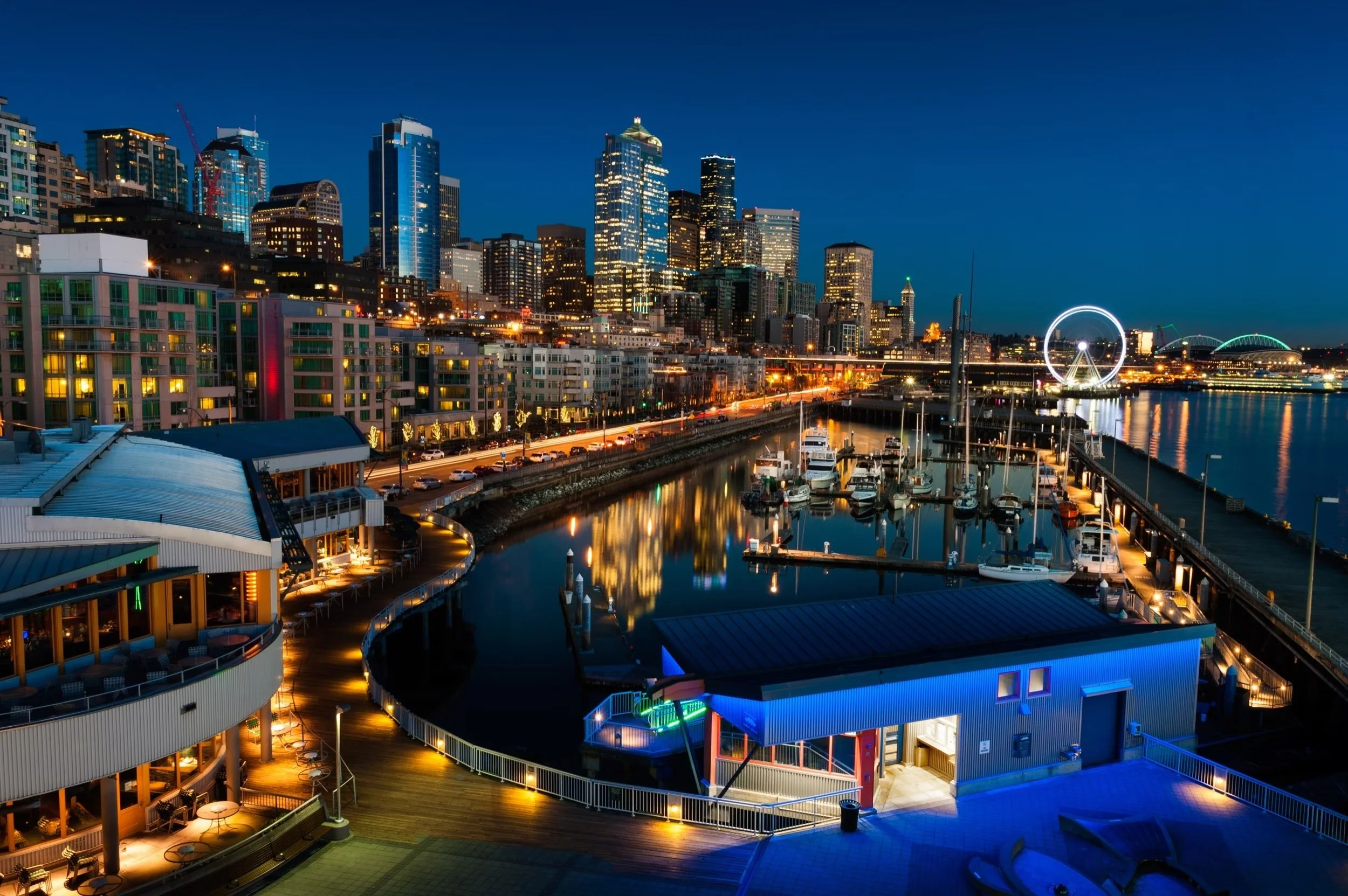 Night view of a city skyline with tall illuminated buildings, a marina with docked boats, a lit Ferris wheel, and a waterfront promenade.