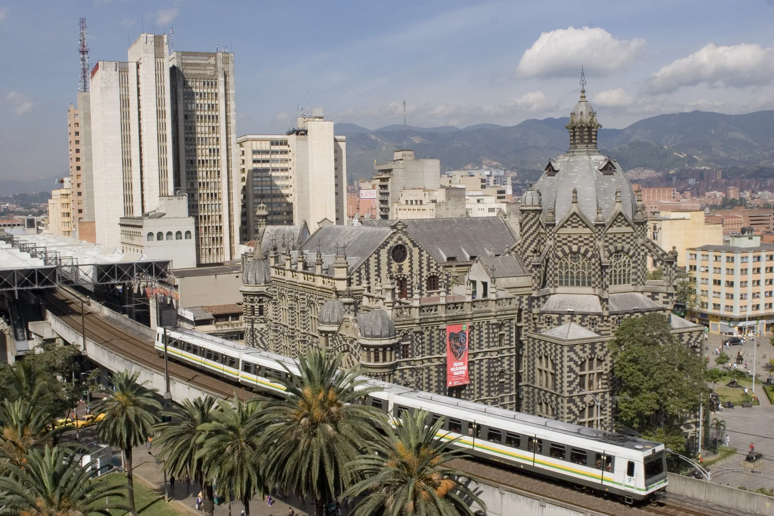 Cityscape with tall modern buildings, a historic church with a black-and-white checkered facade, and a train traveling on elevated tracks surrounded by palm trees.