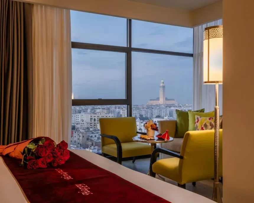 Hotel room with a view of a city skyline and a lighthouse through large windows, decorated with yellow chairs, pillows, and a bouquet of red roses on the bed.