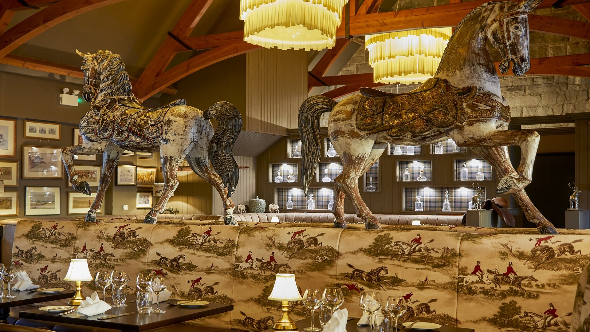 A dining area with two large wooden horse sculptures on a cushioned banquette, framed pictures on the wall, and a wooden ceiling with hanging light fixtures.