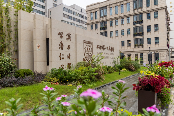 Entrance of Shanxi General Hospital with a stone wall displaying hospital name and logo, surrounded by flowerbeds with colorful flowers and greenery, in an urban setting with tall buildings in the background.