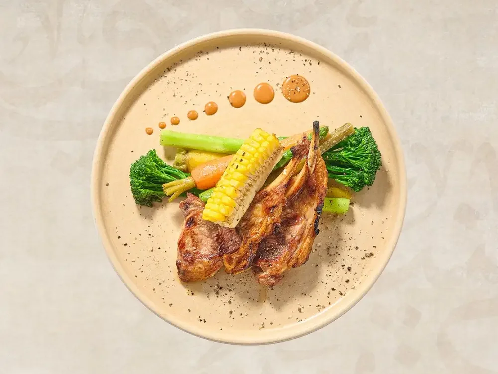Grilled lamb chops with corn on the cob, broccoli, carrots, and green beans on a round beige plate, garnished with a drizzle of sauce and black pepper.