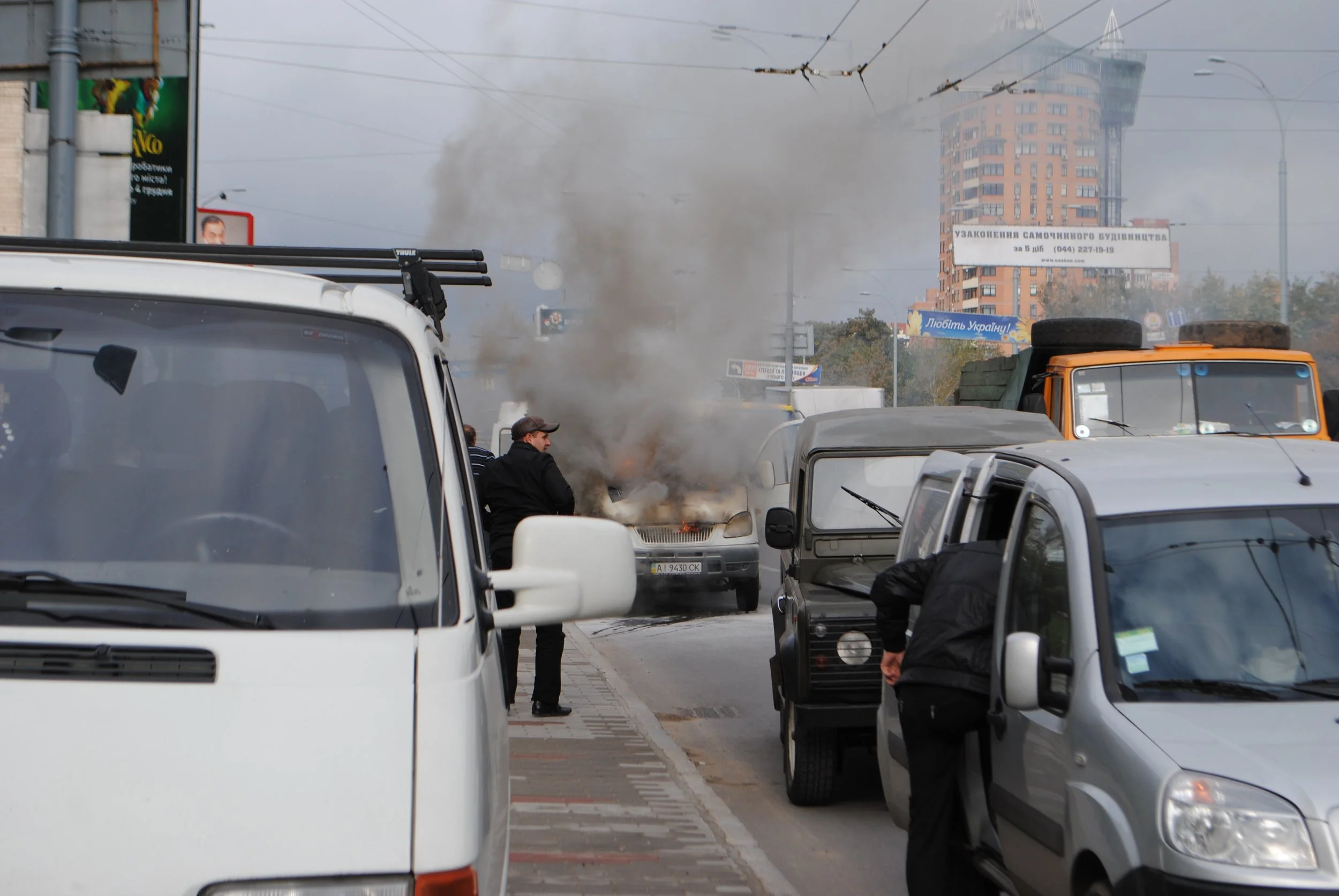 A busy city street with several parked cars and smoke coming from a vehicle in the middle of the road. People are standing nearby. In the background, there is a tall, modern building and various billboards.