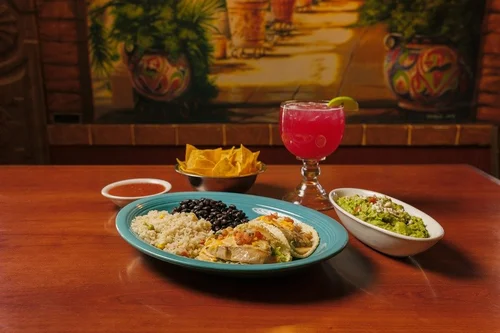 A Mexican meal with three tacos on a turquoise plate, black beans, Mexican rice, guacamole, tortilla chips with salsa, and a pink cocktail with lime on a wooden table.