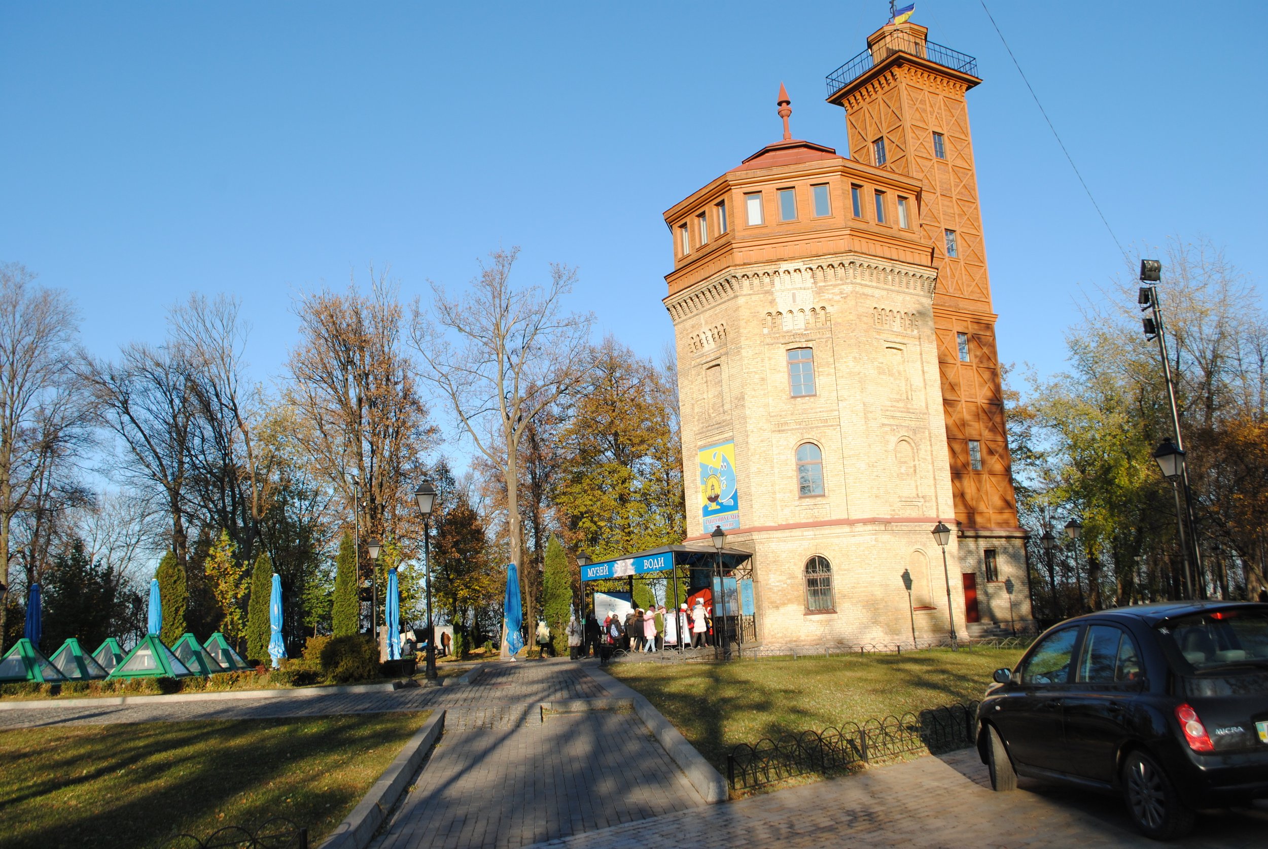 Historical tower with brick and wooden sections, surrounded by trees and a park, with people walking and a parking lot in front.
