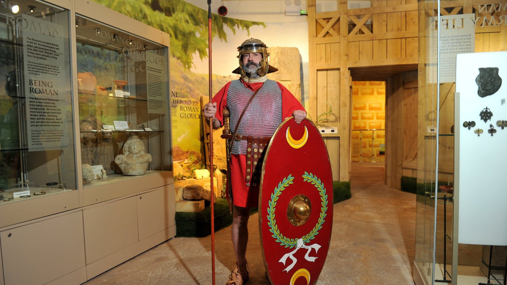 Man dressed as a Roman soldier with a helmet, chainmail, red tunic, shield, and spear standing inside a museum exhibit with displays of artifacts and informational signs.