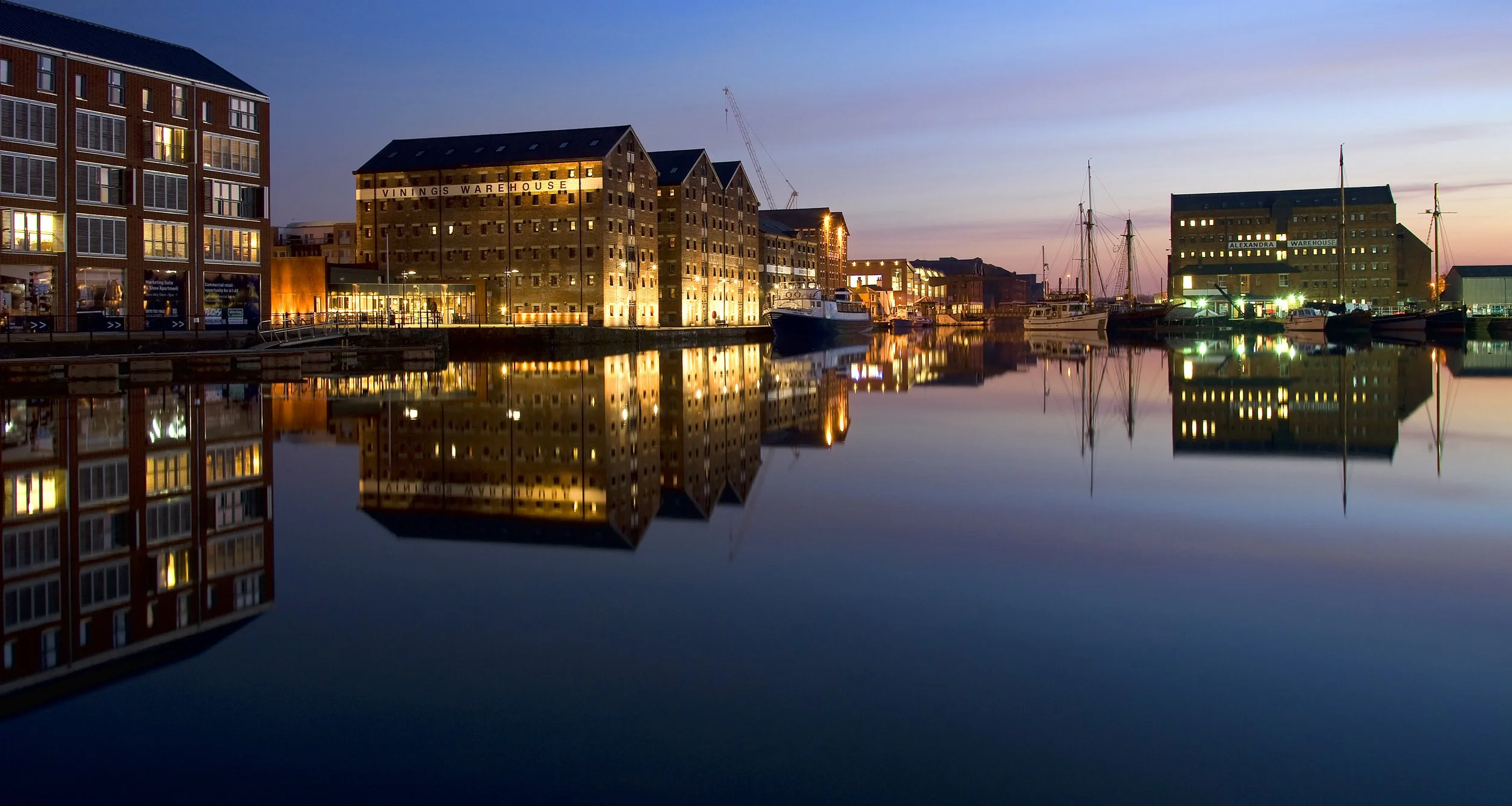 A waterfront scene at dusk featuring lit historic warehouse buildings reflecting in calm water with boats docked along the pier.