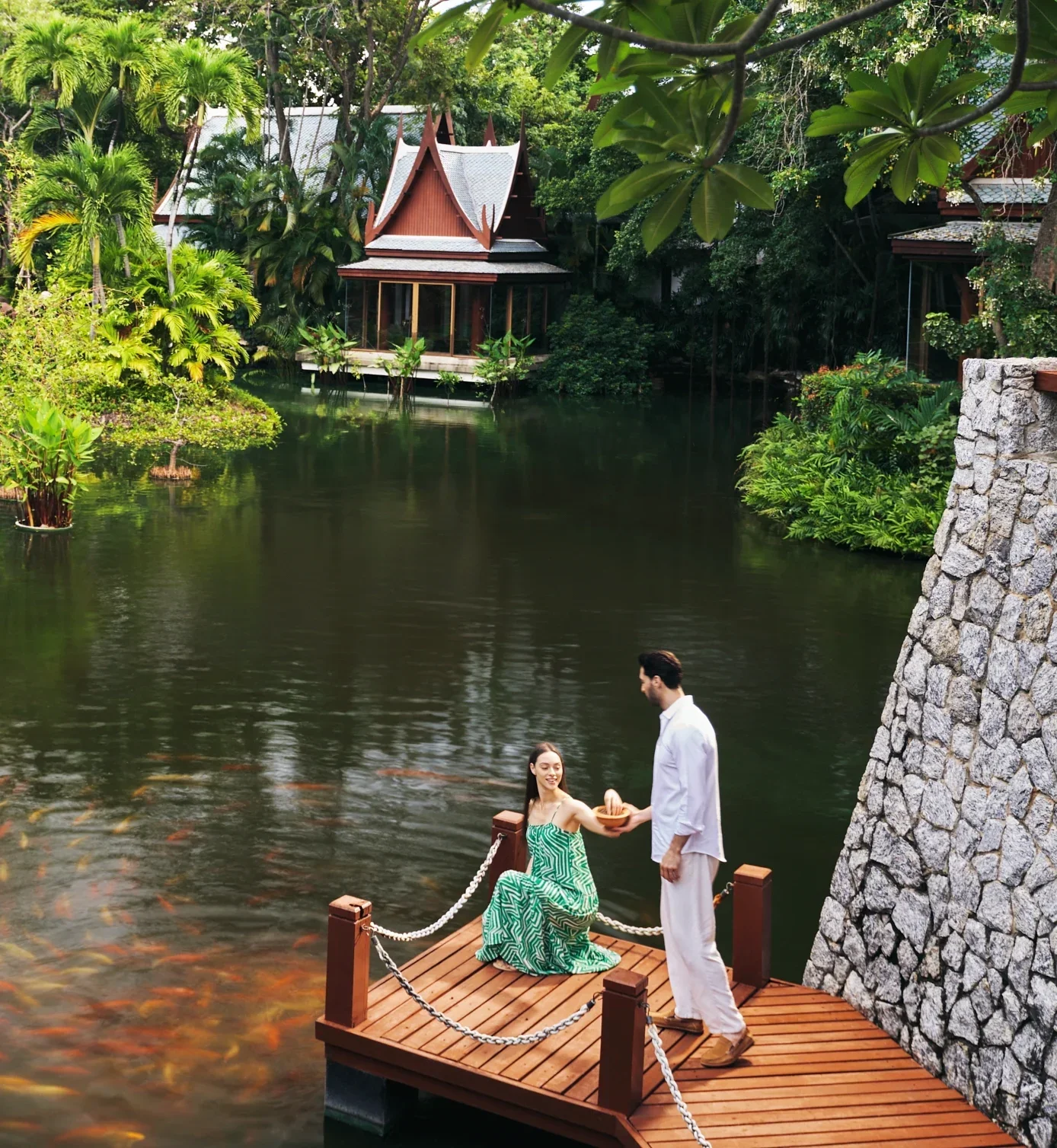 A man proposing to a woman on a small wooden dock by a pond. The woman is seated, holding a wooden bowl, while the man stands in front of her. The pond is surrounded by lush green vegetation and traditional architecture in the background.