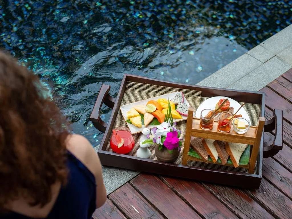 A tray of snacks and beverages on a wooden dock near a pool, including assorted fruit, sandwiches, jams, and a small flower arrangement.