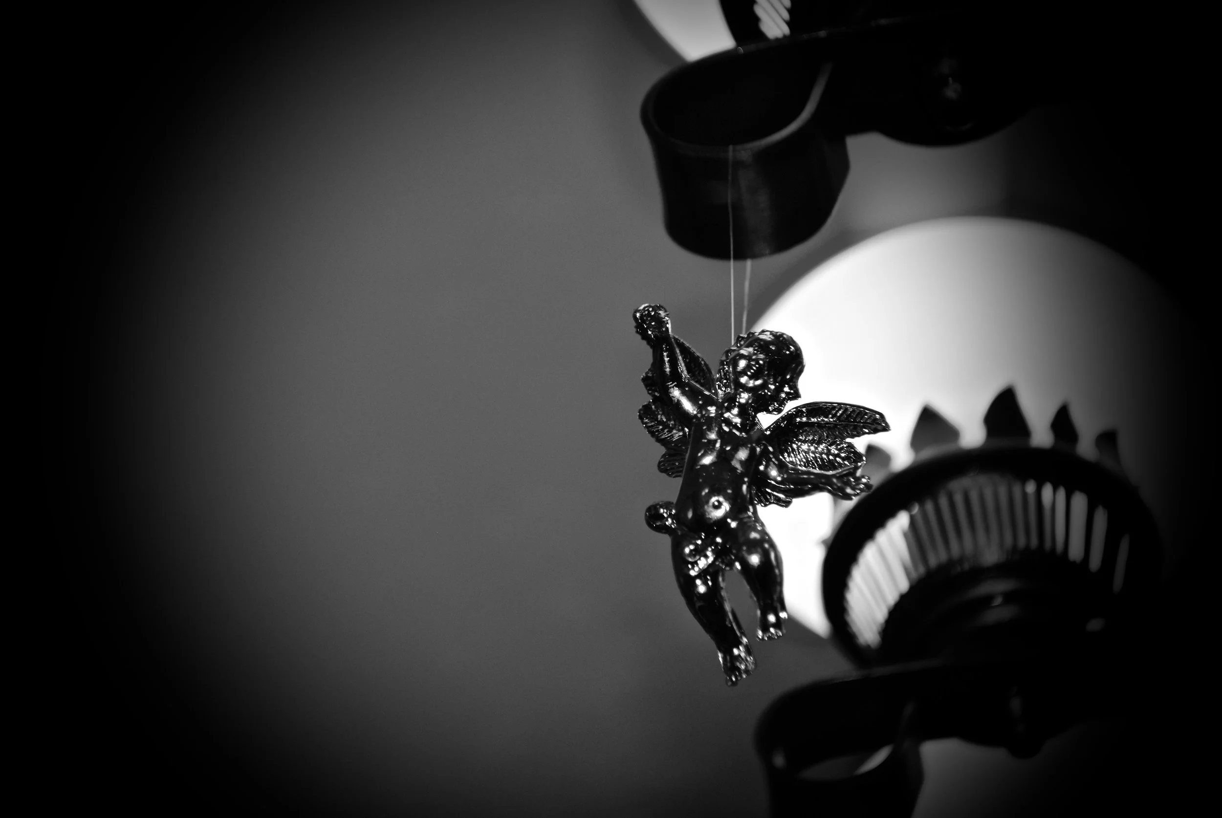A black and white photo of a metallic angel ornament hanging from a structure, with the background out of focus and dark.