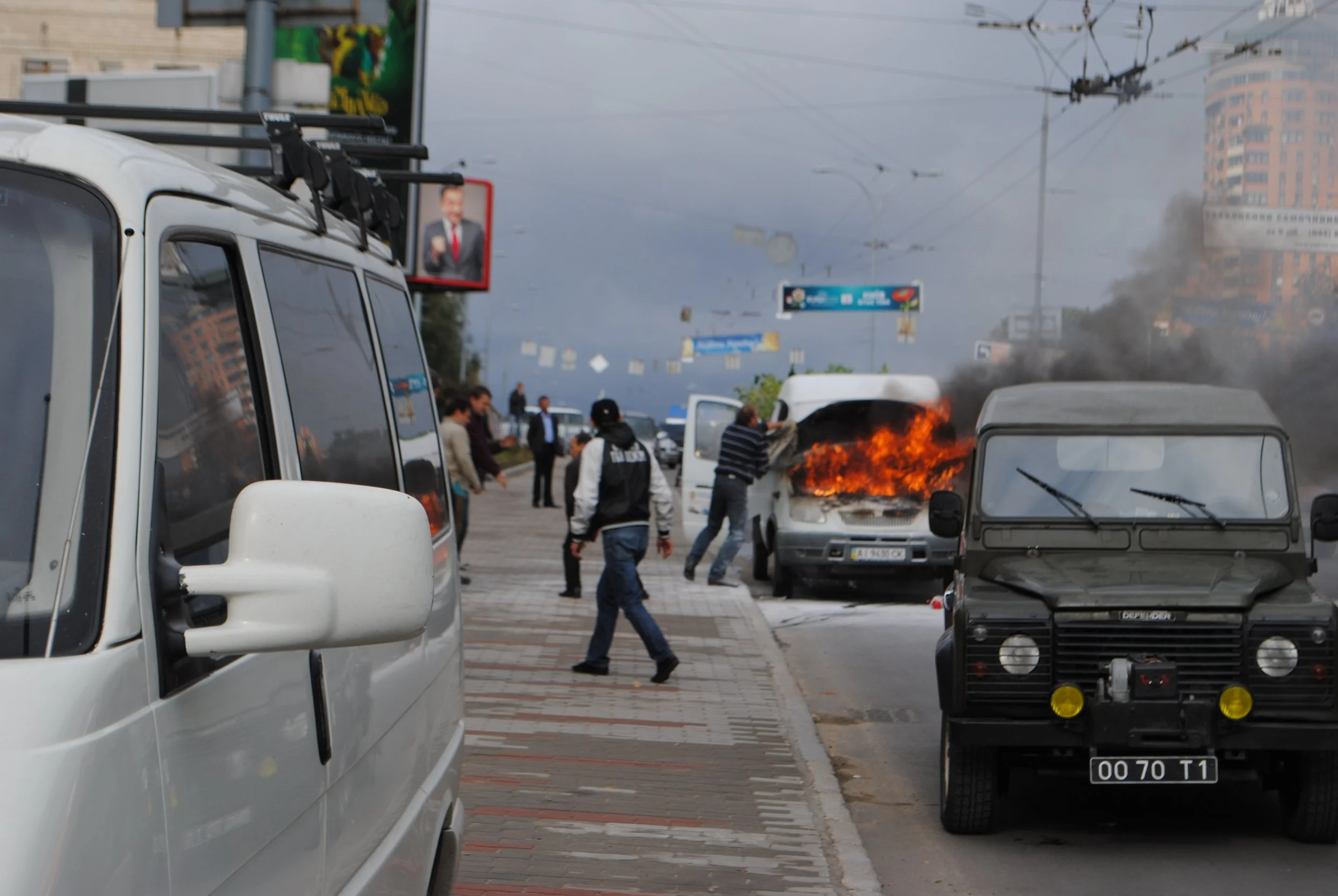 A city street scene showing several vehicles, including a white van and a black van, with one of the vehicles on fire and emitting black smoke. Several people are gathered around, some observing the incident. Overhead, there are electrical wires and 