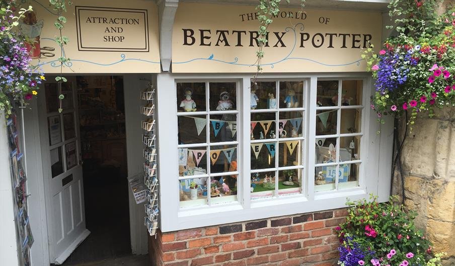 Exterior of a shop called The World of Beatrix Potter, decorated with colorful flowers and hanging baskets, with a display window showcasing Beatrix Potter items and promotional banners.