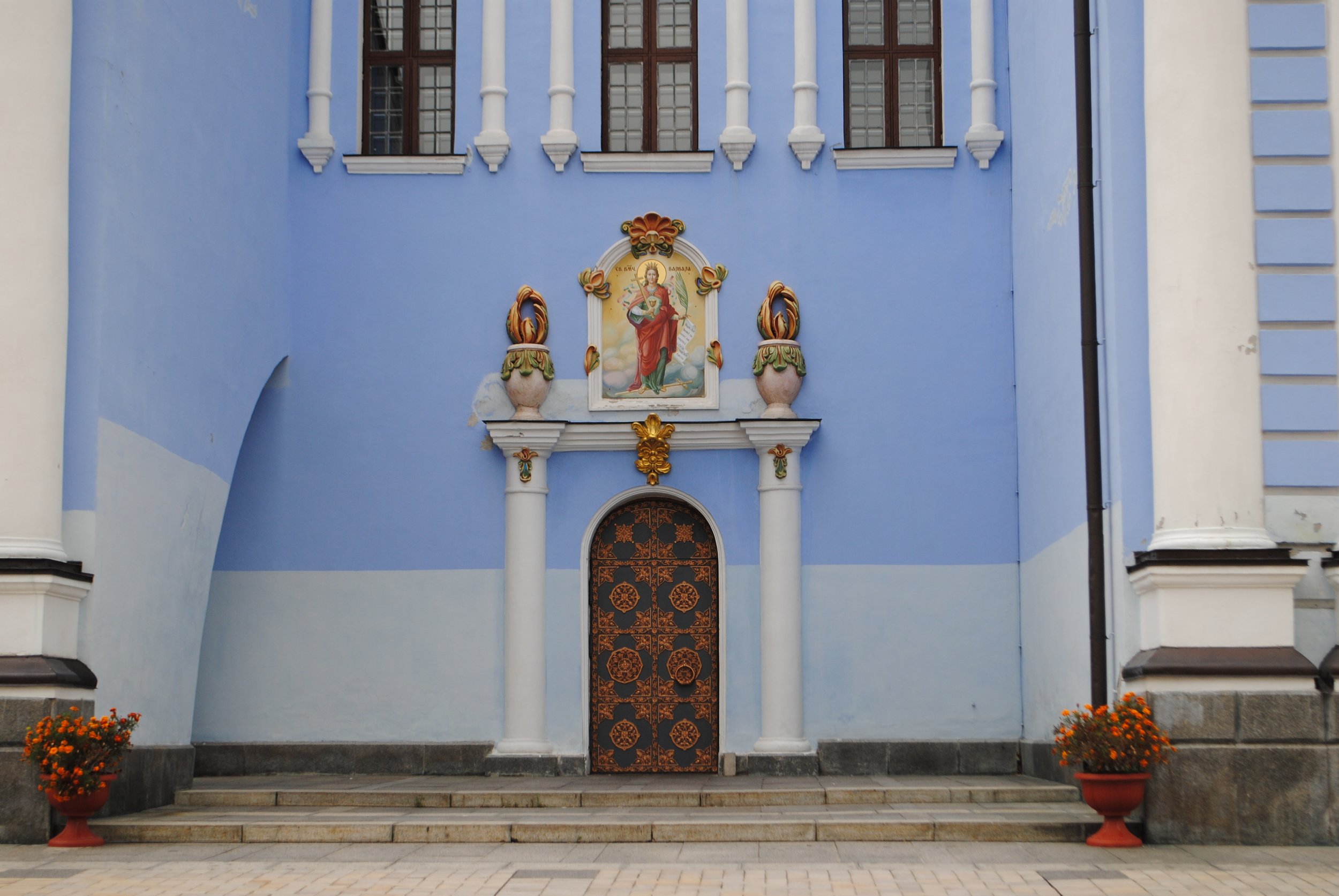 The image shows the front entrance of a church with a blue facade, ornate wooden doors, and religious artwork above the door. Two flowerpots with marigolds are on either side of the entrance, and steps lead up to the door.
