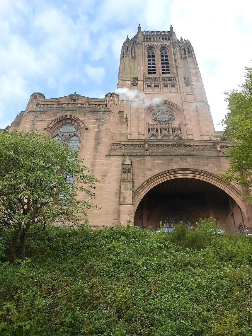 A tall, historic cathedral with a large arched entrance, intricate stonework, and a tower, set against a blue sky with some clouds and green trees and bushes in the foreground.