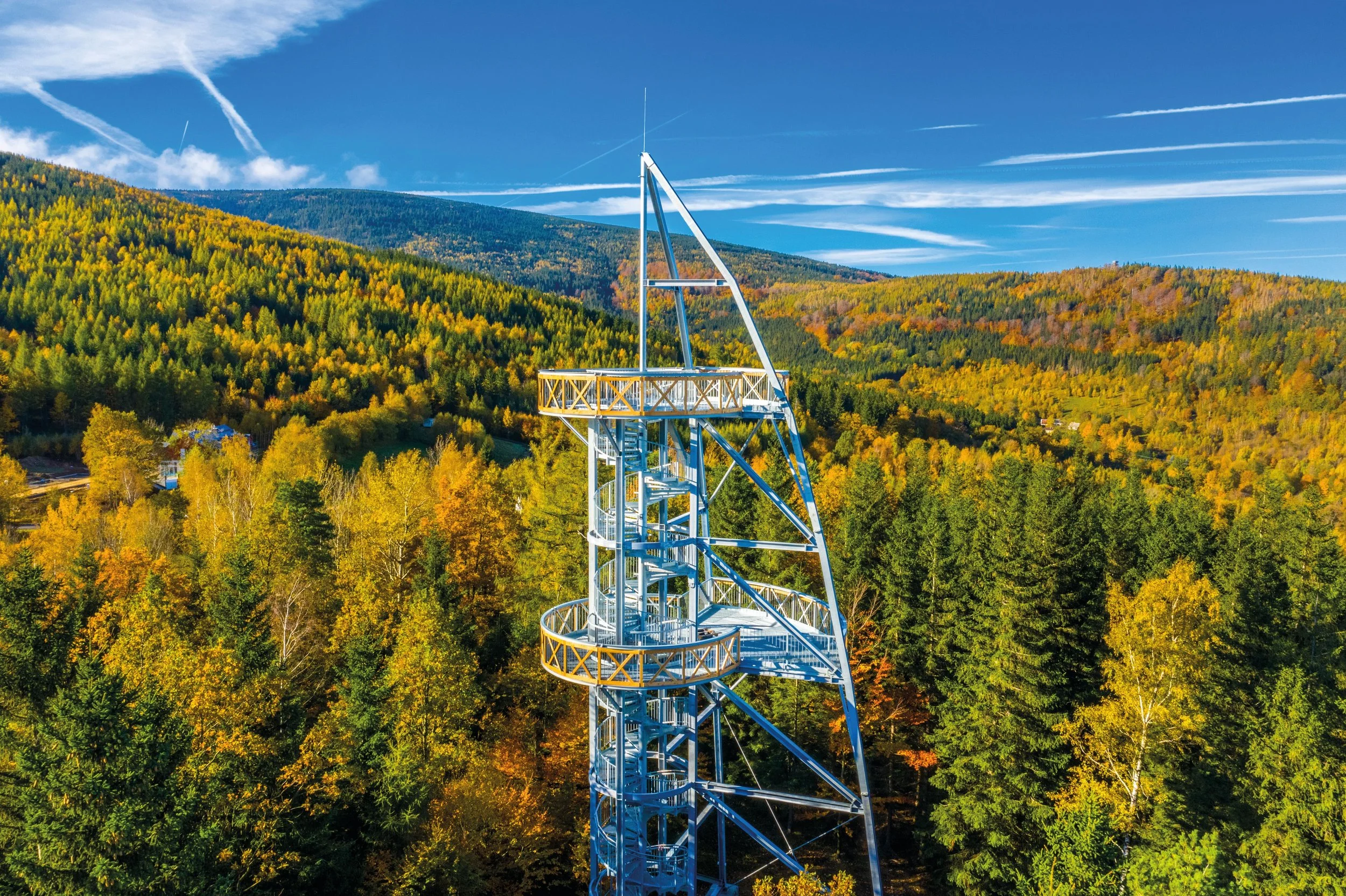 A tall metal observation tower in a forest with trees displaying fall foliage, overlooking distant mountains under a blue sky with scattered clouds and contrails.