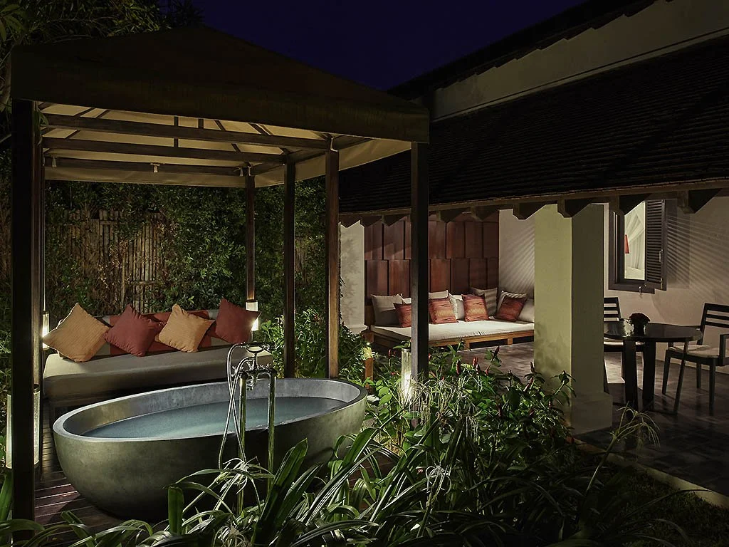 Outdoor patio area at night with a round bathtub, cushioned lounge chairs, and a small dining table with chairs, surrounded by greenery and enclosed by a fence and building walls.