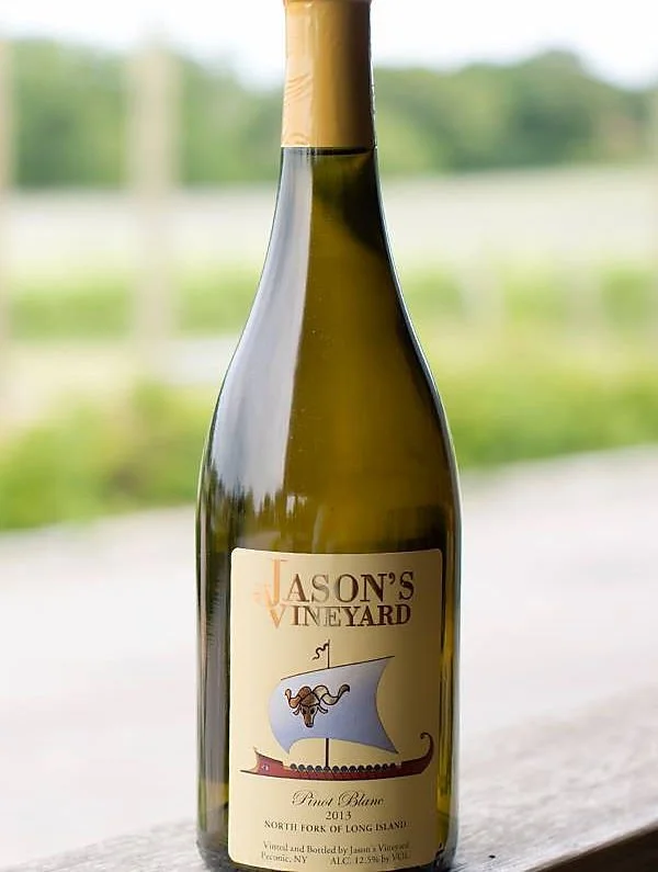 A bottle of Jason's Vineyard Pinot Blanc wine from 2013, North Fork of Long Island, with a label featuring a stylized boat with a sail and a mermaid figure, set against a blurred outdoor background.