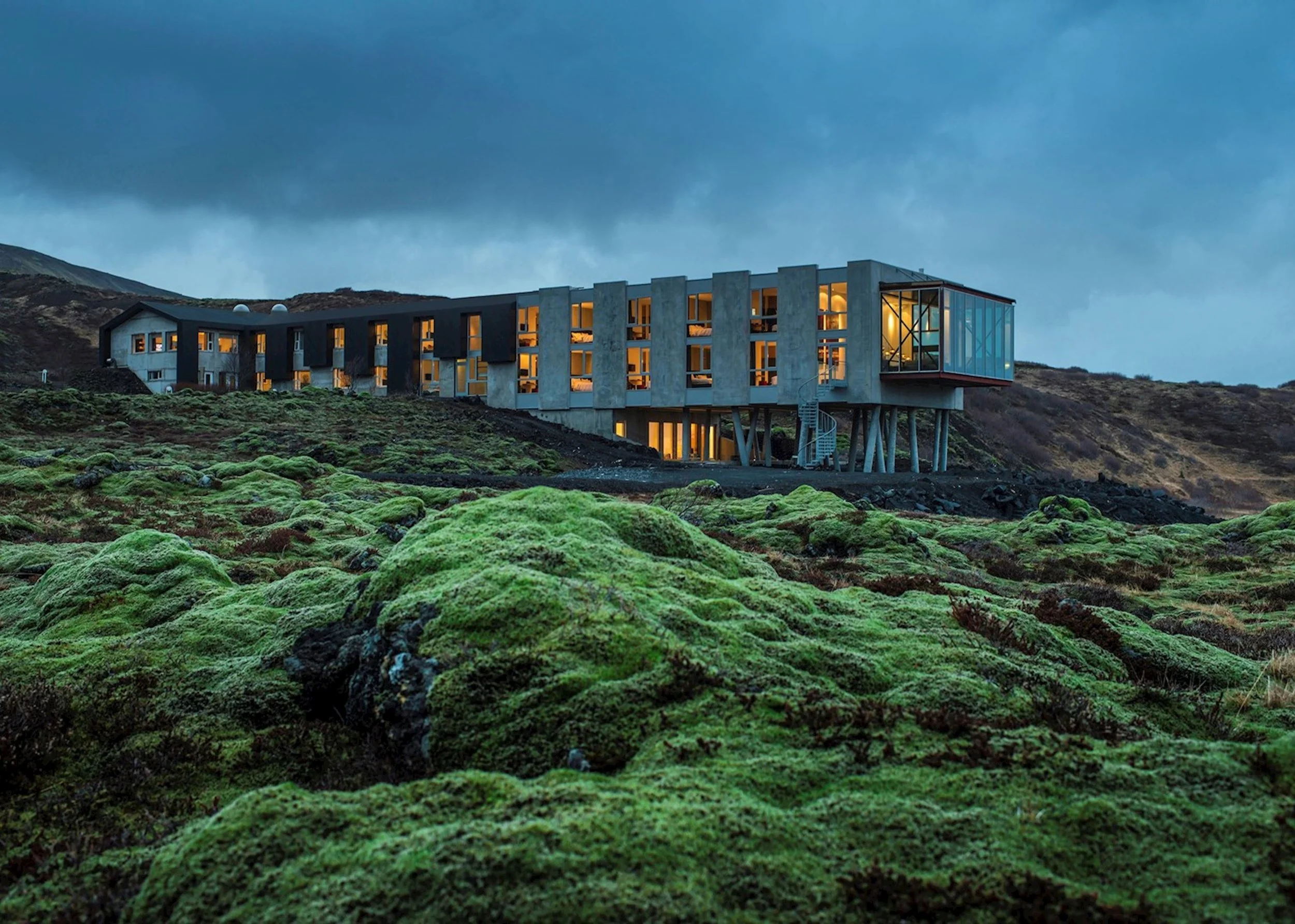 Modern building on a hillside during dusk with green moss-covered foreground and dark cloudy sky.