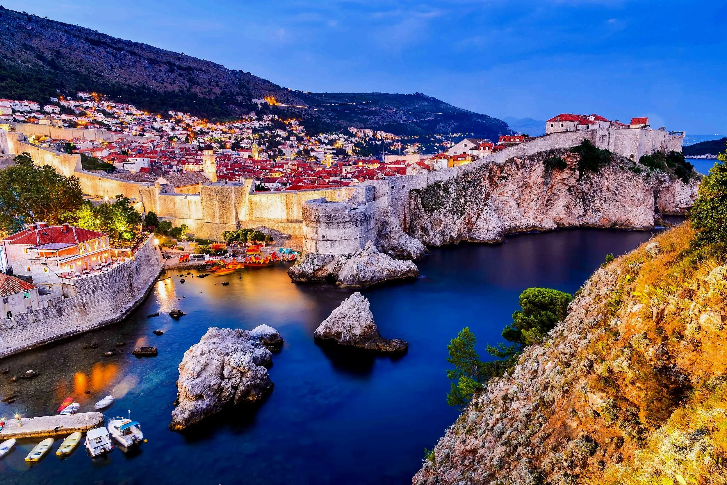A scenic view of the historic city of Dubrovnik, Croatia at dusk, with ancient stone walls, red-tiled roofs, and a calm harbor filled with boats.