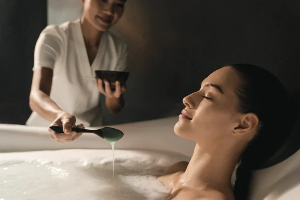 A woman relaxing in a bath with her eyes closed, while a spa attendant pours a liquid into the bath from a ladle.