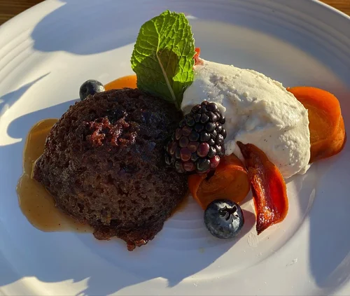Chocolate dessert served with whipped cream, fresh blueberries, a blackberry, a mint leaf, slices of peach, and crispy bacon on a white plate.