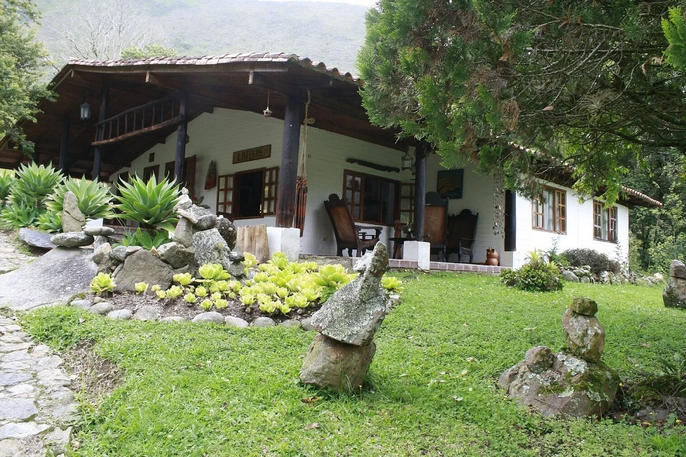 A rustic house surrounded by green grass, plants, and rocks, with a porch and outdoor furniture under a large tree.