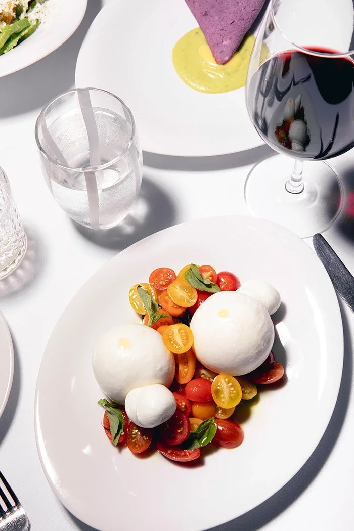 Caprese salad with cherry tomatoes, fresh mozzarella, basil leaves, served on a white plate. Glass of red wine, water, and a purple appetizer on a white plate are also visible on the table.