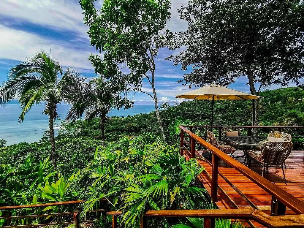 A wooden deck with outdoor furniture and a yellow patio umbrella overlooking a lush, green tropical forest and ocean view under a partly cloudy sky.