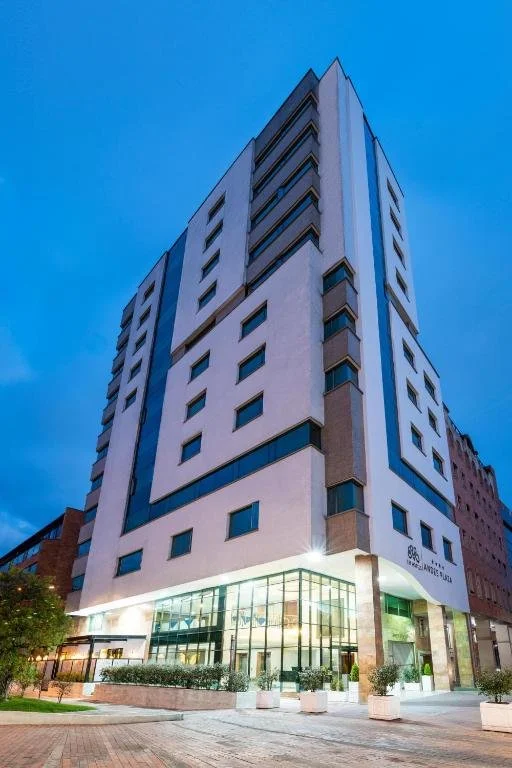 Tall modern multi-story building with a geometric facade, glass entrance, and outdoor plants, against a twilight sky.