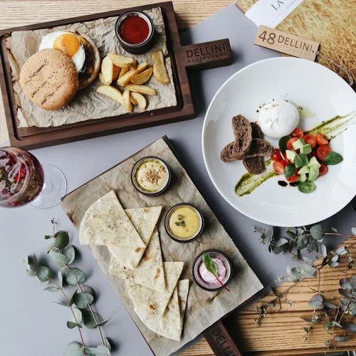 A breakfast spread featuring a burger with a fried egg and fries, a plate with salad, mozzarella, and bread, and a tray of flatbread with dips, along with a glass of water with fruit, on a table decorated with eucalyptus leaves.