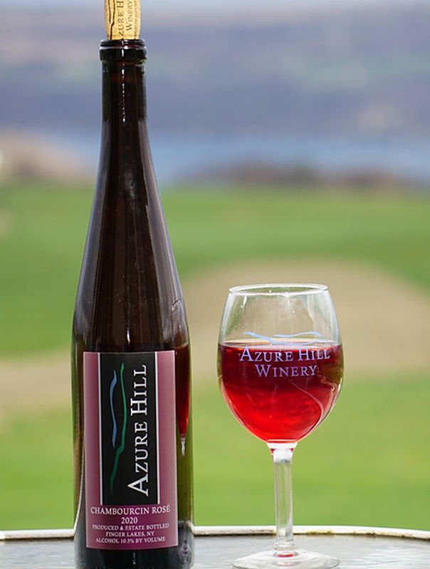 A bottle of Azure Hill Chambourcin Rosé 2020 wine and a filled wine glass with red wine on a table outdoors, with a scenic green landscape and blurred horizon in the background.