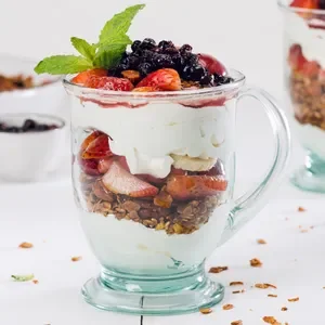 A glass mug filled with layered strawberry, blueberry, and whipped cream dessert, topped with a mint leaf.