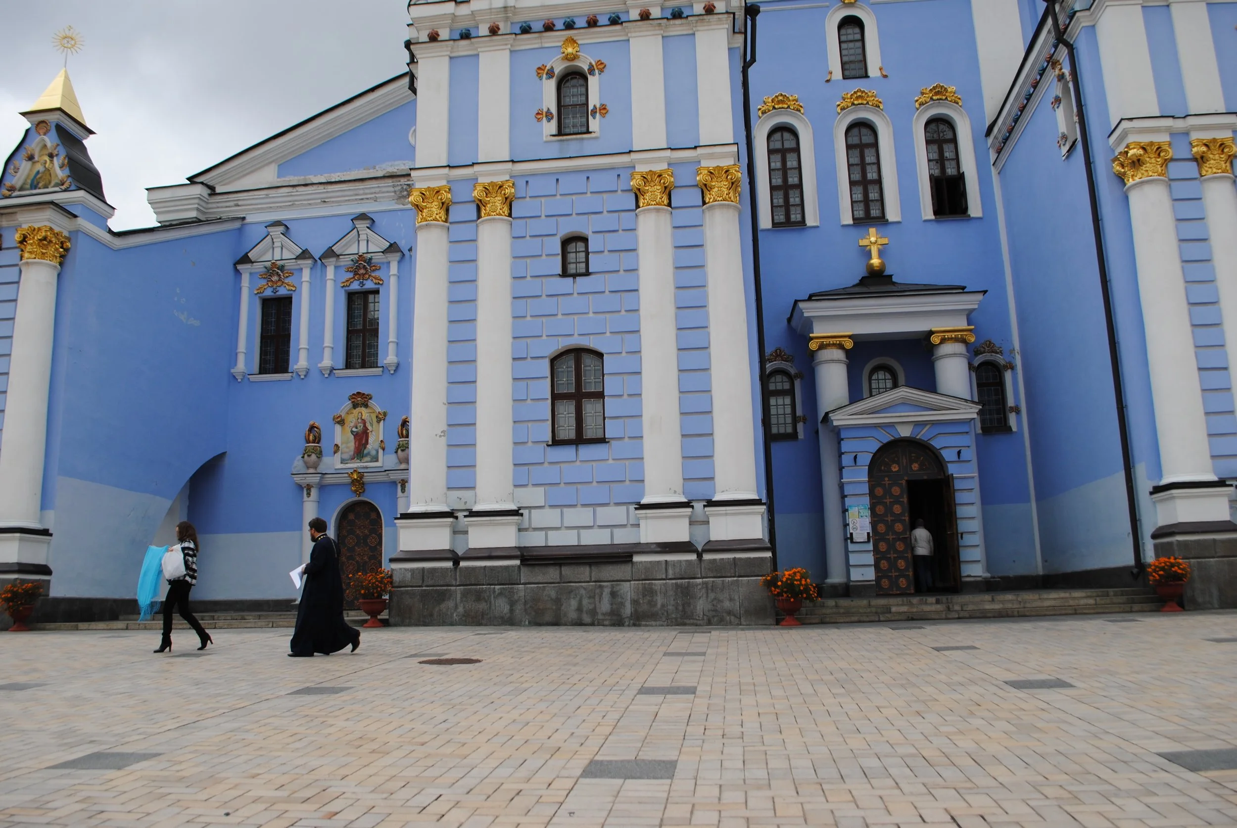 Photo of a blue and white ornate church with gold accents, multiple arched windows, and a cross on top. Several people are walking in front of the church on a paved area with orange flowers in pots nearby.