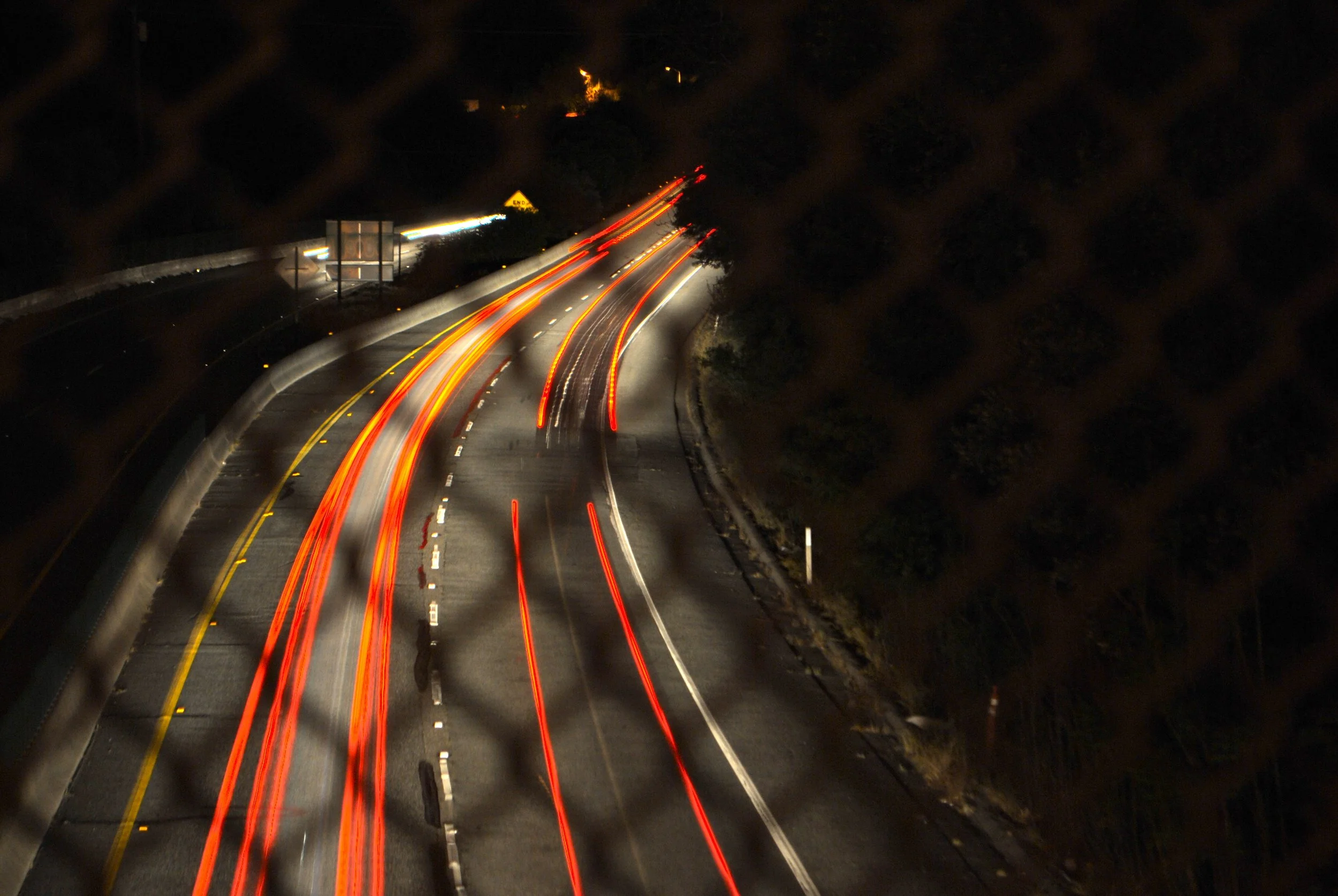 Long exposure photo of a highway at night, with streaks of red and white light from vehicle taillights and headlights, and a chain-link fence in the foreground.