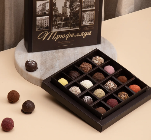 A box of assorted chocolate truffles with various flavors and toppings on a beige surface, with three truffles outside the box and a black and white photo album with Russian writing in the background.