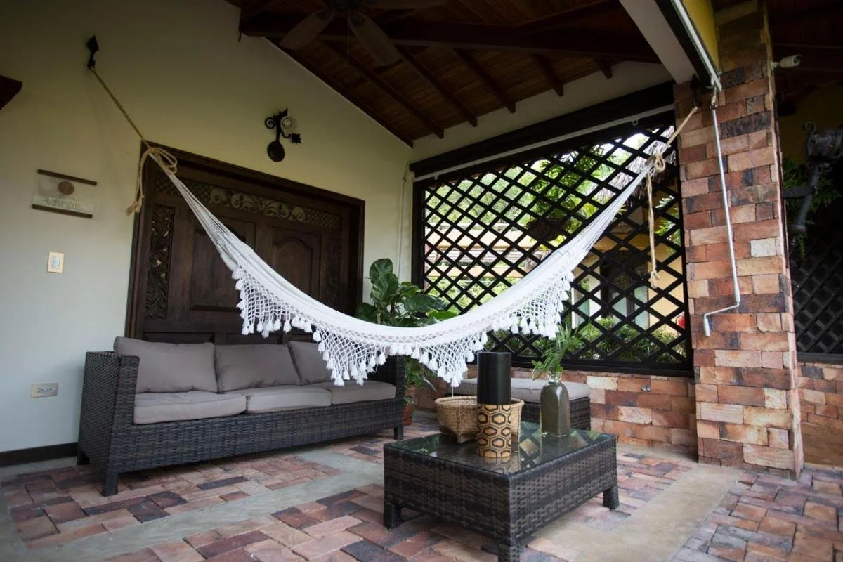 Outdoor patio with wicker sofa, coffee table, potted plants, and a white hanging hammock with fringe, brick flooring, and lattice privacy screen.