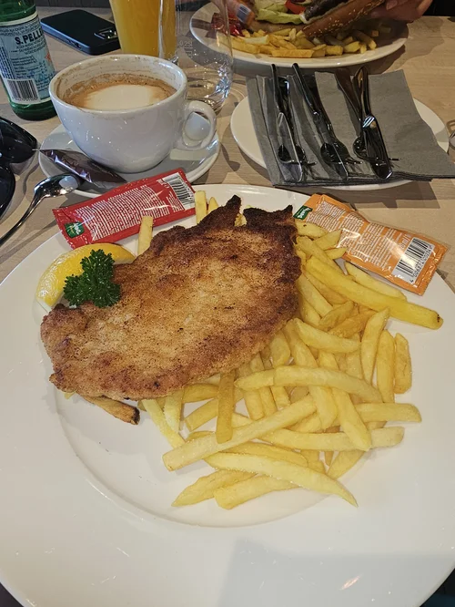 Plate with breaded fried fish fillet, French fries, lemon wedge, and parsley. In the background, coffee, soda, and a plate with salad and fries.
