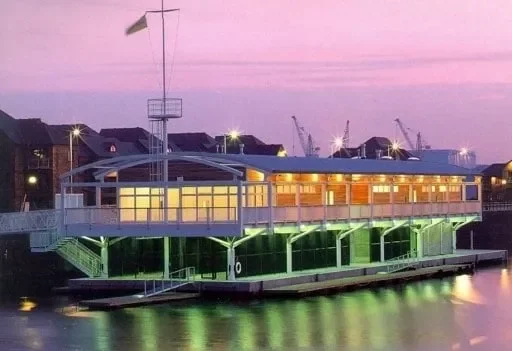 A floating house on a body of water with large windows illuminated at sunset.