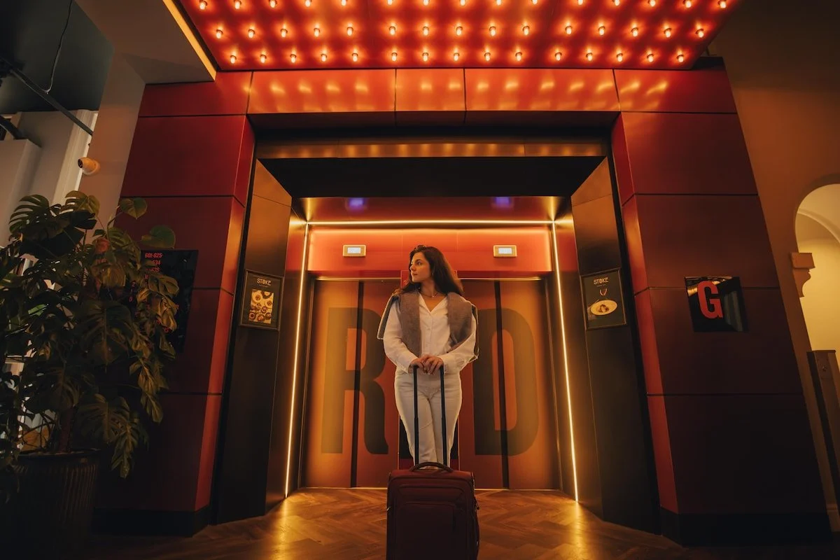 A woman with brown hair wearing white clothes holding a rolling suitcase in front of a brightly lit elevator with orange and black decor.