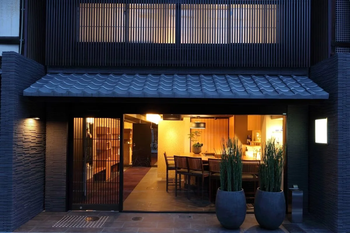 The entrance of a modern home with black sliding glass doors, a tiled roof, two large potted plants outside, and warm interior lighting visible through the glass.