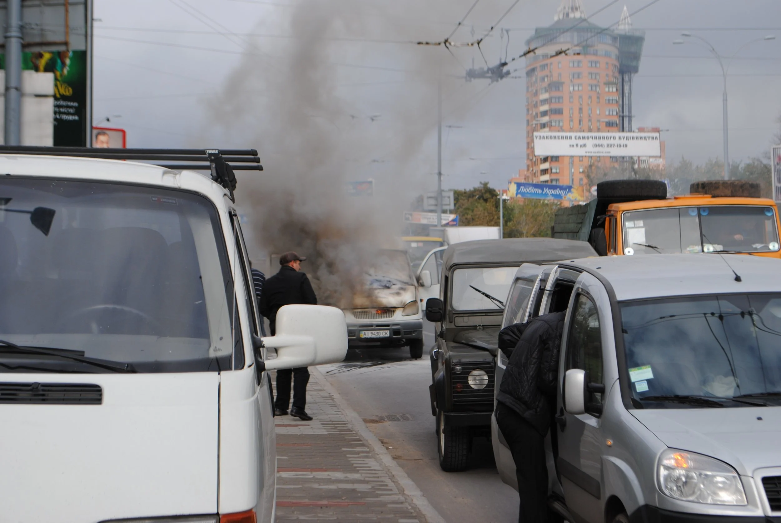 Street scene with vehicles, people, and smoke in the air, in an urban area with a tall building and billboards in the background. Kiev November 2011, Marina Parrish ©