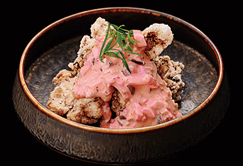 A dark bowl containing pieces of fried chicken topped with pink sauce and garnished with a sprig of greenery.