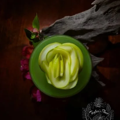 Close-up of a green cup filled with sliced green apple pieces, with pink flowers and a piece of driftwood in the background.